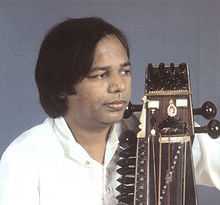 A middle-aged man wears a shirt and looks to the side with a bowed instrument held close to his body.