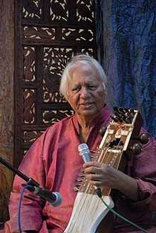 An old man sitting in front of an ornamented partition wall speaks into a microphone and holds a bowed instrument.