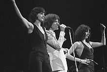 Three people stand onstage next to each other singing. The man in the middle wears white clothes and on either side is a woman wearing black.