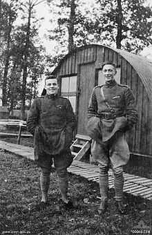 Two men in military uniform standing side-by-side. The man on the left if short and stocky, while the man on the right is tall and thin. In the background is a cabin.