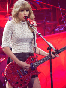 A blonde-haired woman holding a sequined red guitar, singing on a stage lit in red and violet