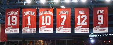 The banners of six retired numbers.  The banners, from left to right, read "Yzerman 19" "Sawchuk 1" "Delvecchio 10" "Lindsay 7" "Abel 12" "Howe 9". The Yzerman banner has a small "C" at the top right corner.
