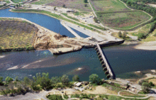 View from the air of a river flowing through the center from right to left; a dam blocks the water flow in the right half presumably to send some of the water to a canal that leads off to the upper left.