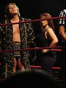 A man and a woman standing in a wrestling ring with red ropes. The man is standing on the left, and has light brown hair. He is wearing a long black coat with gold detailing, short black wrestling tights, and has a wrestling championship around his waist. The woman is standing beside him, but has her face obscured by the ring ropes. She has shoulder length brown hair, and is wearing a black top and black trousers.