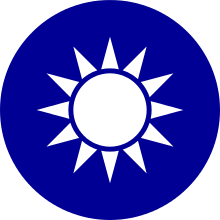 A blue circular emblem on which sits a white sun composed of a circle surrounded by 12 rays.