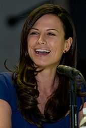 A seated woman with brown hair extending below her shoulders and wearing a blue shirt smiles as she looks to her right.