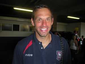 A man in a navy blue open-necked shirt with a crest and the word "mitre" on the chest, smiling.