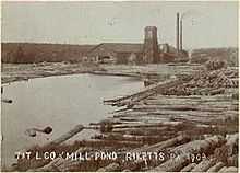 Sepia-tone photo of a pond surrounded by large logs. At the far end of the pond is a large building with a square tower and two smokestacks. Label is "T&T L CO MILL POND RICKETTS PA 1903" (i.e. Trexler and Turrell Lumber Company Mill Pond ...)