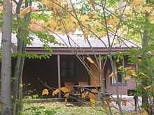 Photo of a brown clapboard-sided cabin with porch, framed by the branches and green, yellow, and orange leaves of small trees.