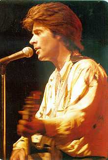 A young man in profile playing a guitar and standing before a microphone.