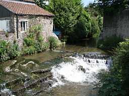 Water flowing through a channel and over a weir between a building and a wall. Vegetation on both sides of the water.