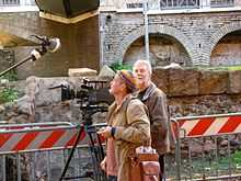 A photograph of two men with a black video camera on a black tripod standing in front of a grey-bricked building with rounded arches