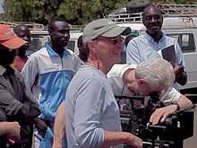 A photograph of a group of standing men, the one in the foreground smiling and wearing a green baseball cap, glasses, and a grey shirt