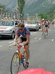 A cyclist going towards the camera, with other cyclists and team cars behind him, and mountains in the background.