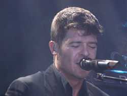 A picture of a brown-haired man from the shoulders up. He is wearing a black shirt and singing into a microphone.