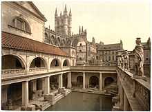 A late-nineteenth century Photochrom of the Great Bath at the Roman Baths. Pillars tower over the water, and the spires of Bath Abbey – restored in the early sixteenth century – are visible in the background.