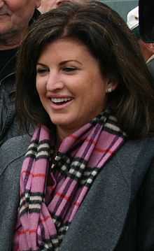 A photograph of a woman facing left and looking left while wearing a pink, black, and white plaid scarf, a grey jacket, and earrings