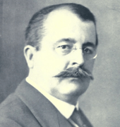  Head and shoulders of a man with receding hair, rimless glasses and a heavy moustache. Formally dressed with collar and tie, he is facing right but his eyes are turned to the camera.