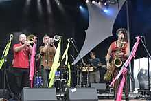 From left to right on a stage: a trombonist, trumpeter, two drummers in the background, and a saxophonist, all playing in bright colors