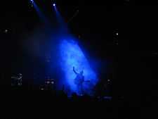 A man in a smoky blue spotlight singing at a microphone stand, holding a guitar in one hand and raising the other hand; off to his side and not in the light, another man standing and playing an electric keyboard