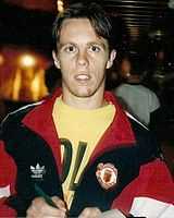 A photograph of a man with short brown hair. He is wearing a black tracksuit top with red trim over a yellow shirt.