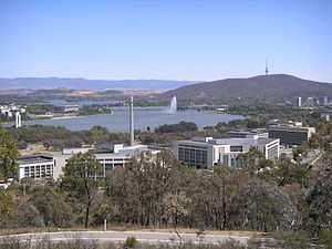 A group of multi-story office buildings. A lake, mountains and a jet of water are visible in the background.