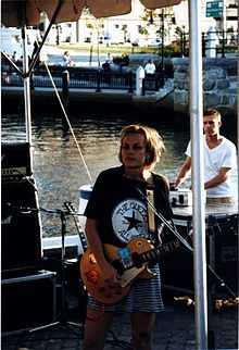 Ruthie Morris standing onstage with a guitar, wearing a black t-shirt with a modified Chuck Taylor All-Stars logo