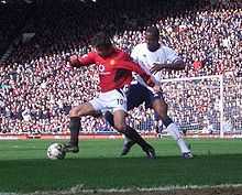 A photograph of two men playing football. The man on the left, who is wearing a red shirt, white shorts and black socks, is shielding the ball from the man on the right, who is wearing a white shirt, navy blue shorts and white socks with navy blue trim.