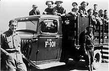 Black and white image of a stakebed truck from the 1930s with two men standing in front and about a dozen men in the back. The truck is labeled ECW (Emergency Conservation Work, original name of the CCC) above the windshield, and "S-118 F-101" on the door.