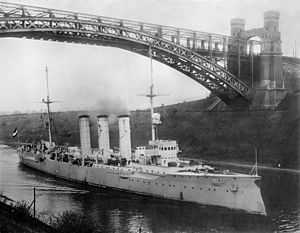 A large ship with three smokestacks passes under a tall arch bridge