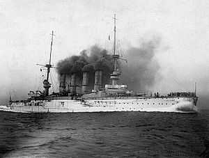 A large white warship belching thick black smoke plows through the water