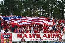 American fans, dressed in red, cheer in bleachers as they hold a large American flag over themselves at a soccer match.