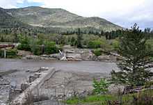 A muddy river flows through a gap between the ruins of a dam on either shore. A wooded hill or mountain rises in the background.