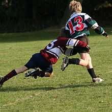A child running away from camera in green and black hooped rugby jersey is in the process of being tackled around the hips and legs by an opponent.