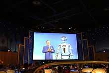 A photograph of a projection screen depicting a man with short, grey hair wearing a necktie while standing next to a grey, physically anthropomorphic robot