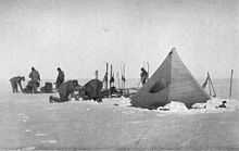  Six men are working with sleds and camping equipment, close to a pointed tent pitched on a snowy surface. Nearby, upright skis have been parked in the snow