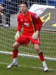 A young man, wearing a red top, shorts and socks, and large, white gloves. He is standing on a white line, painted onto a grass field. A net is visible behind him.