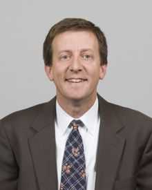 A portrait shot of Scott S. Harris, looking straight ahead. He has short brown hair and is wearing a charcoal blazer with a blue patterned tie over a white collared shirt.