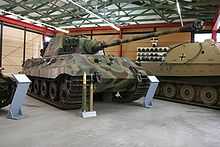 A three quarters view of a large tank with a flat-faced turret, dull yellow, green and brown wavy camouflage, on display inside a museum. The frontal armor is sloped. The long gun overhangs the bow by several meters. Two waist-high cartridges sit on their bases in front of it.