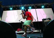 A man wearing a red T-shirt and light green shorts on a stage singing into a wired microphone. A white screen and blue stage lights serve as his backdrop in an evening setting.