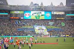 A stand of a stadium with fans holding a large banner depicting the Space Needle. Green and blue flags wave throughout the stand and the flags of different nations are held by people on the field.
