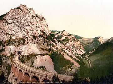 A photochorm picture of the a curving railroad bridge coming out of a tunnel bored into a large mountain.  In the background another bridge and mountains are visible.