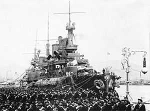 A black and white picture of a ship at anchor with uniformed men to the ship's left side.