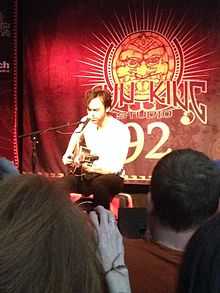 A man seated on a briefcase converted into a make-shift drum playing guitar in front of a microphone