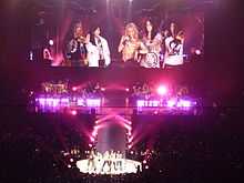 Five ladies are standing in front of a stage and a closeup of them is being projected onto a large screen behind them. There is pink-coloured lighting on the stage.