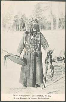 Faded black and white photo of a Mongol Buryat shaman, a masked man wearing a thick robe, an apron covering his chest, and a round hat with covering his face down to his nose. He is holding a drum in his right hand, and two decorated wooden sticks in his left hand. The caption in Russian and French means "Buryat shaman."