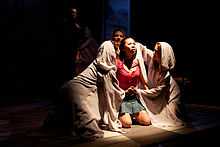 A photograph of a kneeling woman wearing a red shirt and a blue jean skirt surrounded by three other women wearing white cloaks and staring at her