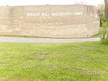 Behind a large area of mown grass stands a beige brick wall which curves around a corner to the right. The wall bears the words "Sheriff Hill Methodist Church", which follow the curve of the wall.