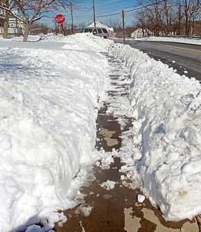 A sidewalk at the bottom of a crudely cut trench through the snow. Some snow on it is melting in the sun.