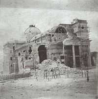 Photograph showing a domed masonry building with its left corner and most of its facade reduced to rubble and only one tower on the right and half of the pedimented porch intact